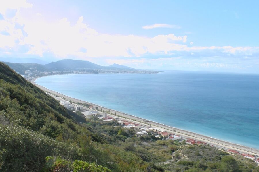 Coastal landscape with lush hills, sandy beach, clear sea, and distant mountains.