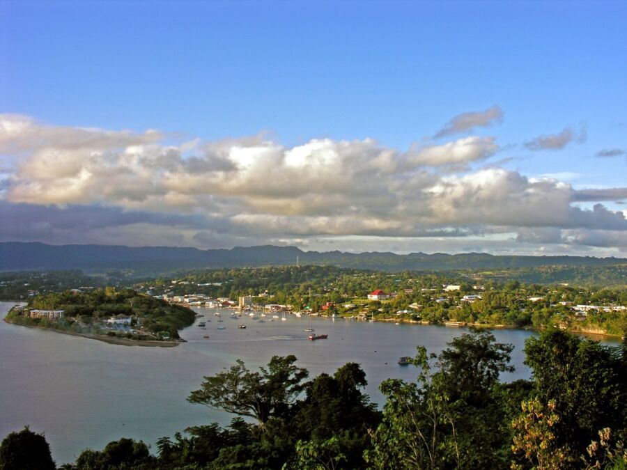 Serene coastal bay view with lush greenery, boats, rolling hills, and scattered clouds.