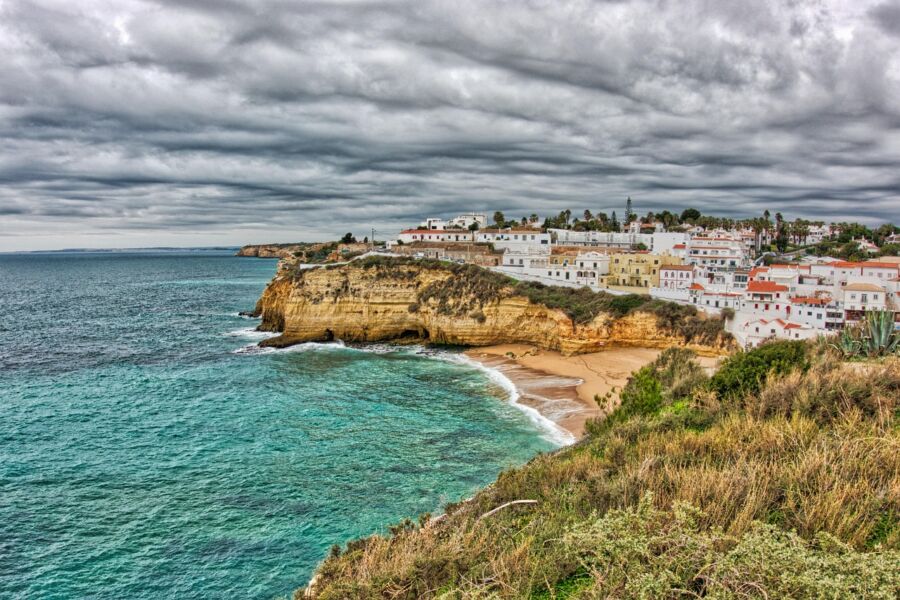 Coastal Carvoeiro cliff, turquoise ocean, white village, terracotta roofs, overcast sky, lush greenery.