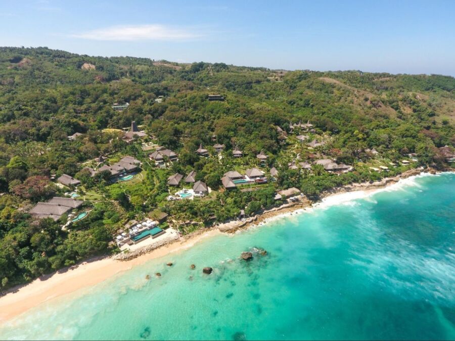 Aerial view of tropical luxury villas by turquoise ocean and lush green hills.