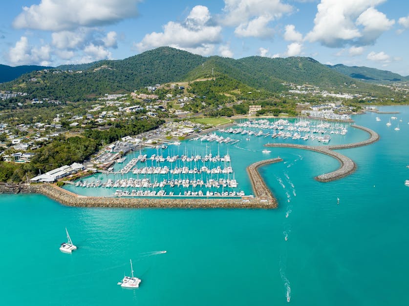 Aerial view of tropical marina with yachts, catamarans, turquoise water and lush coastal hills.