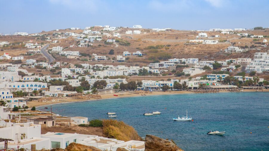 Pristine Mykonos beach with turquoise water, white Cycladic architecture, and terraced hills.