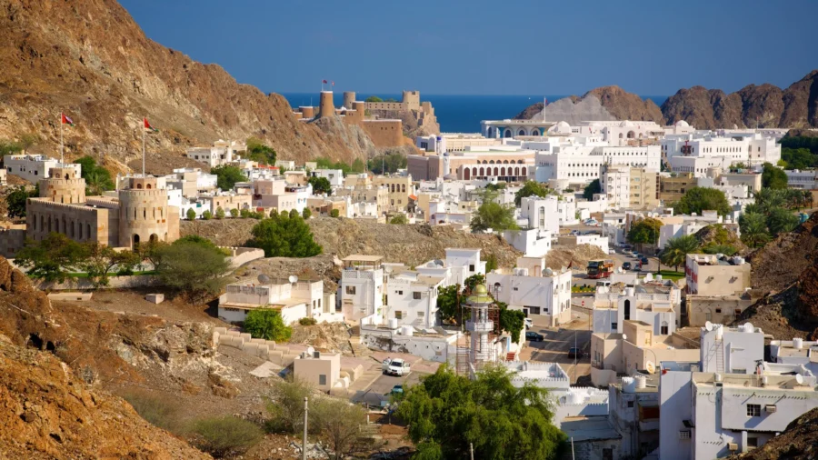 Middle Eastern coastal city with historic forts, mountains, and white buildings overlooking the blue sea.