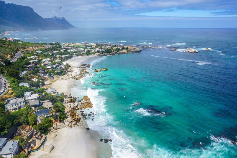 Coastal landscape with ocean, sandy beach, rocks, residential homes, mountains, and vibrant greenery.