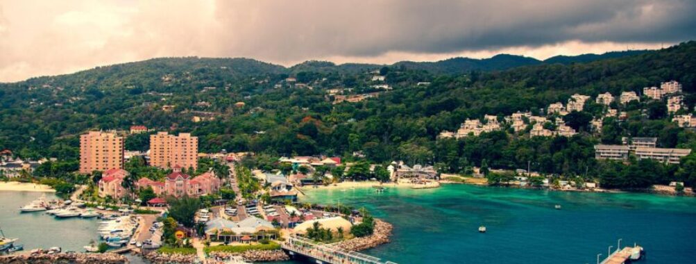 Tropical marina with boats, colorful buildings, lush hills, and sandy beaches under cloudy skies.