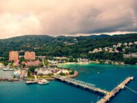 Tropical marina with boats, colorful buildings, lush hills, and sandy beaches under cloudy skies.