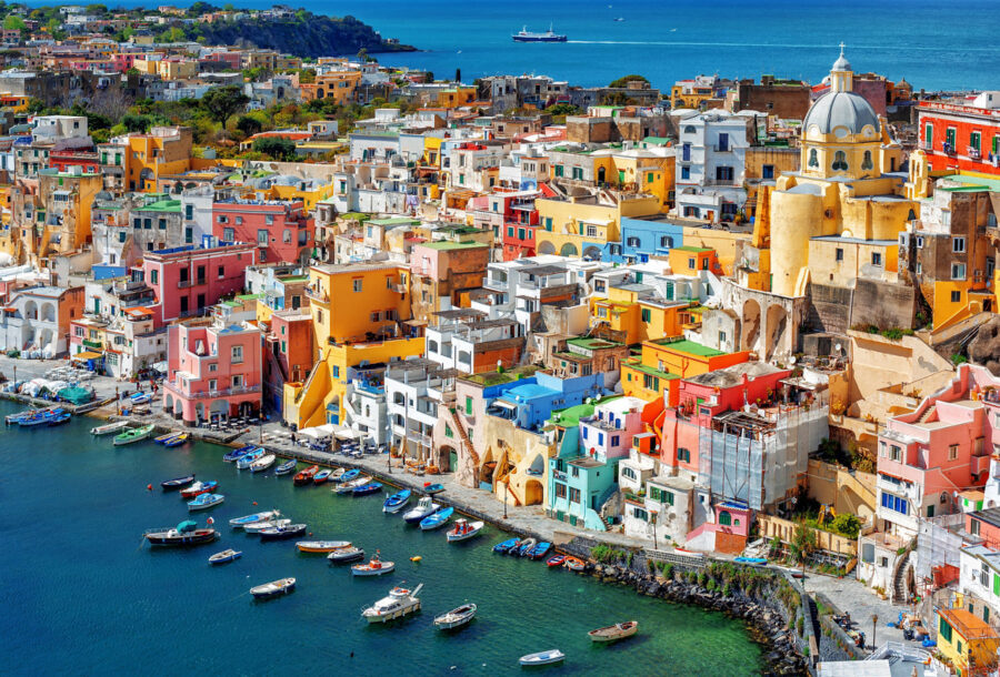 Sunlit pastel coastal harbor of Procida with colorful houses, boats, and Mediterranean sea.