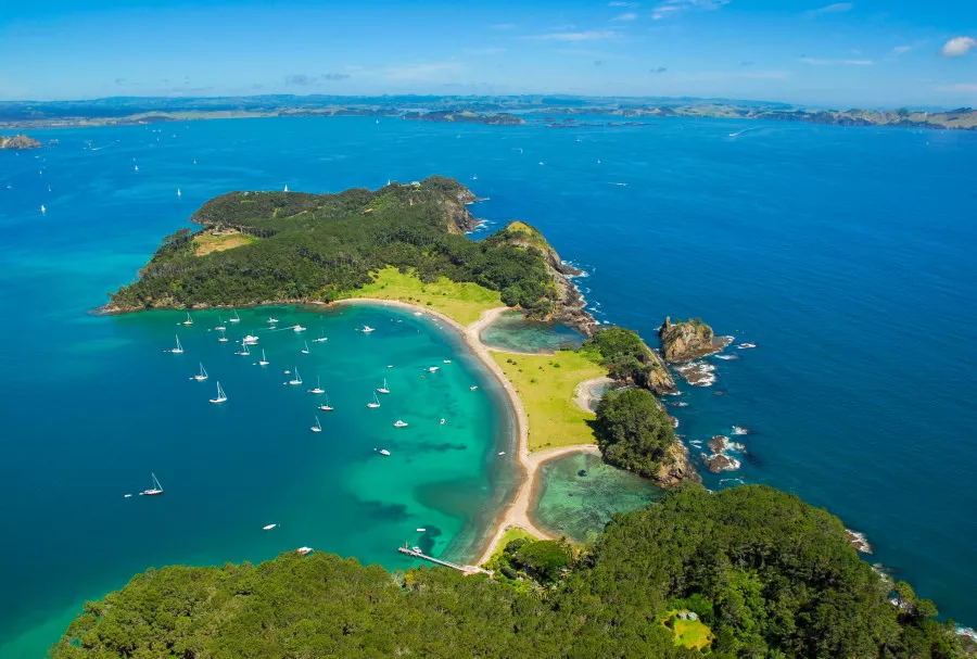 Aerial view of Bay of Islands peninsula with forest, beaches, turquoise water and anchored boats.