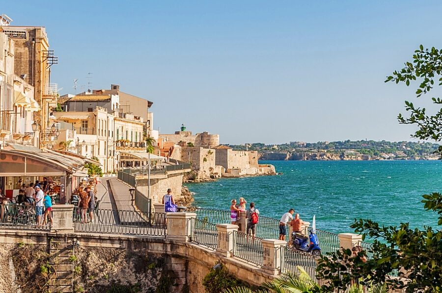 Mediterranean seaside promenade with cafes, historic architecture, people, and scenic ocean view.