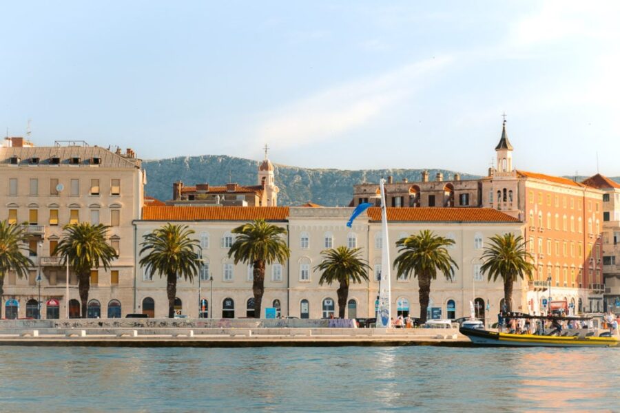 Scenic coastal town with waterfront promenade, historic architecture, palm trees, boats, and hills.