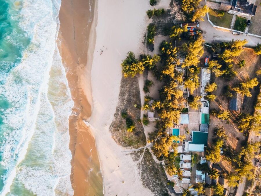 Aerial view of tropical beach resort with turquoise ocean waves, sandy shore, trees, and pools.