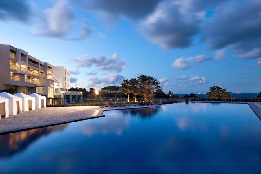 Luxury coastal resort infinity pool at dusk reflecting sky, hotel building, palm trees, and ocean.