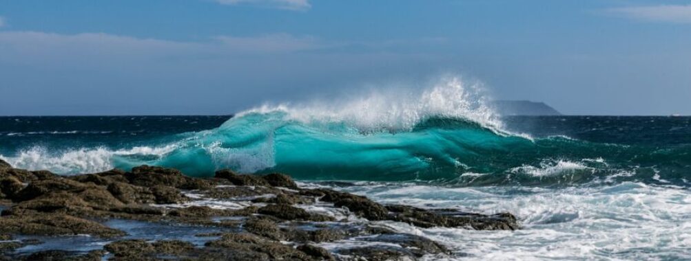Vibrant ocean wave crashing on rocky shore under a sunny, cloud-dotted sky.