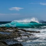 Vibrant ocean wave crashing on rocky shore under a sunny, cloud-dotted sky.