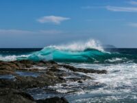 Vibrant ocean wave crashing on rocky shore under a sunny, cloud-dotted sky.