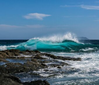 Vibrant ocean wave crashing on rocky shore under a sunny, cloud-dotted sky.