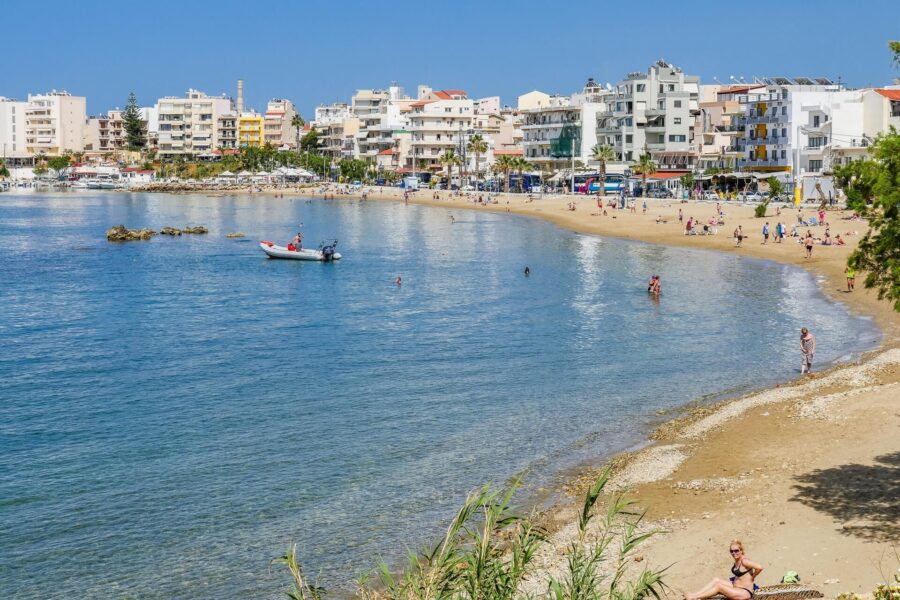 Sunlit beach with turquoise water, lively shoreline, vibrant buildings, and recreational activities.