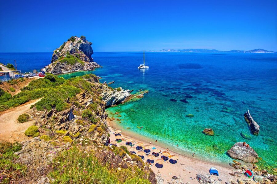 Scenic coastal beach with rocky shoreline, verdant hills, sailboat, and vibrant umbrellas.