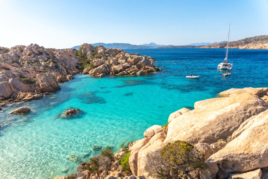 Serene coastal bay with turquoise waters, rugged rocks, sailboat, and distant mountains.