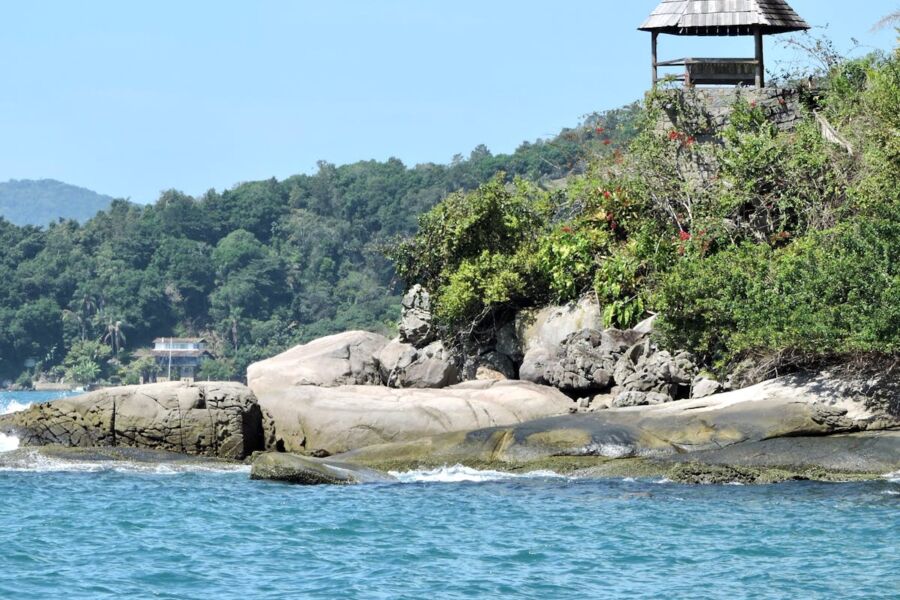 Serene coastal landscape with rocks, turquoise water, lush greenery, and a rustic gazebo.