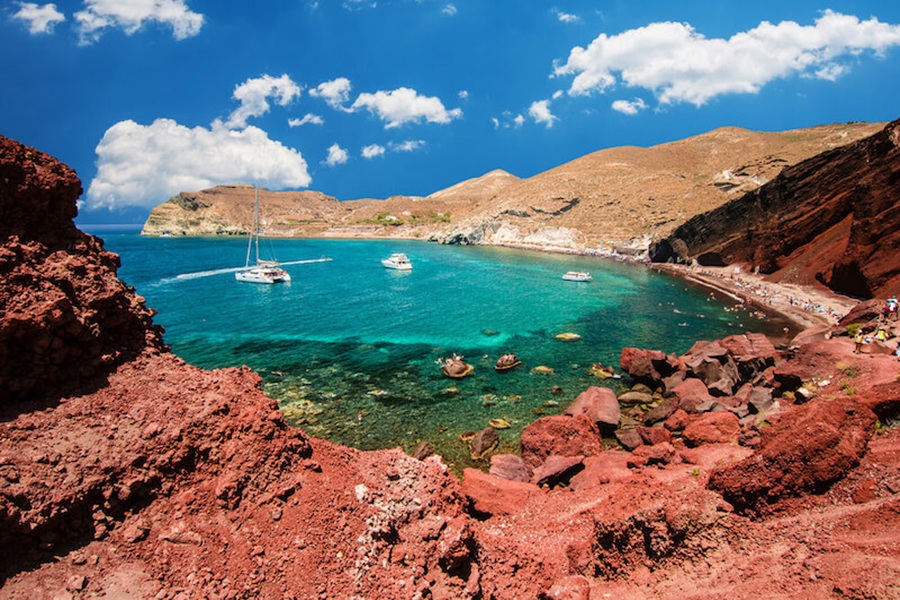 Stunning coastal landscape featuring red rocks, turquoise waters, boats, and scenic cliffs.