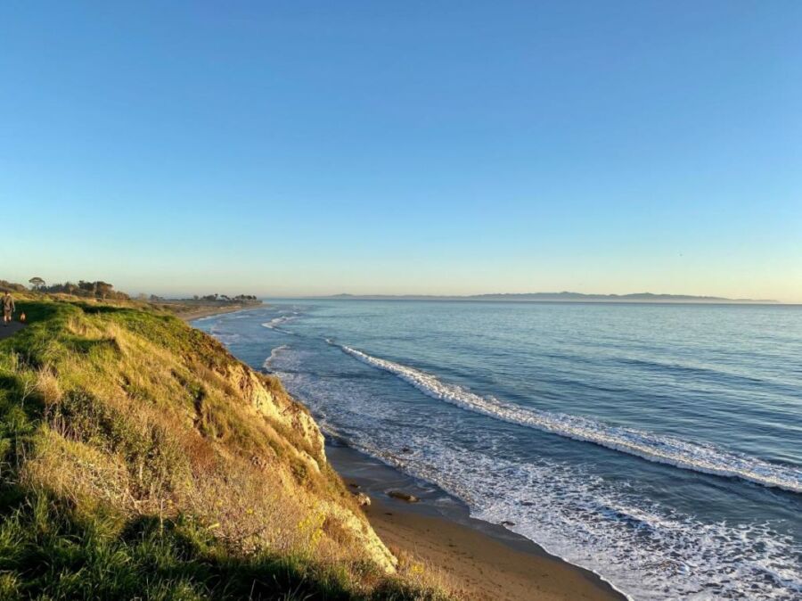 Serene coastal landscape with lush cliffs, sandy beach, and calm ocean under clear blue sky.