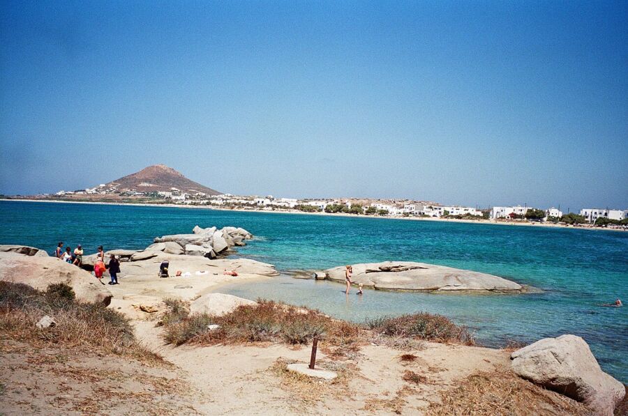 Coastal scene with beachgoers, rocky shore, Mediterranean architecture, vibrant ocean, and sunny sky.