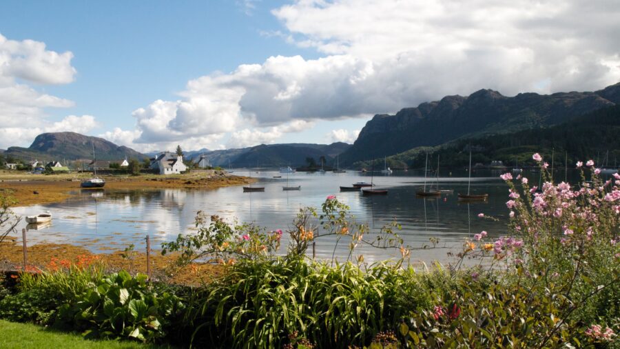 Scenic coastal landscape with boats, flowers, hills, and reflections in tranquil water.