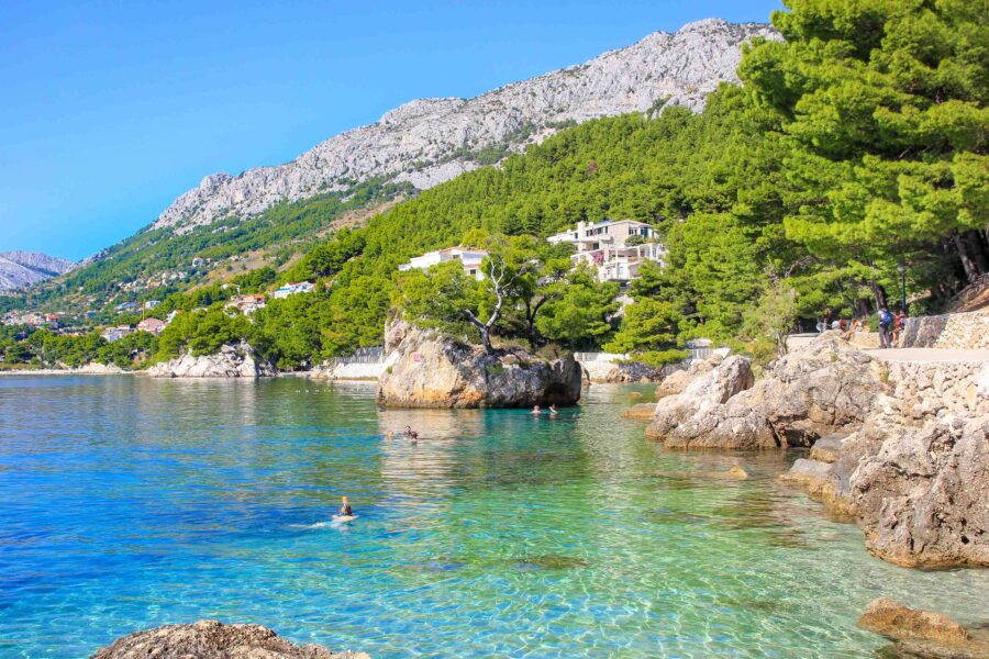 Turquoise sea, rocky coast, lush hillside, and serene beach scene with people enjoying nature.