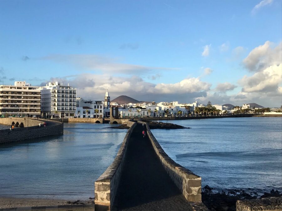 Stone causeway leading to Arrecife coastal town, white buildings, church tower and calm blue sea.