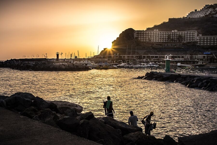Sunset in Puerto Rico, Gran Canaria: serene coastal scene with boats, people, and rocky backdrop.