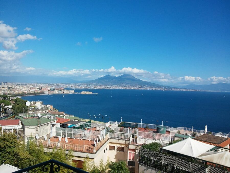 Lungomare Caracciolo terrace overlooking Naples bay, rooftops, blue sea, and Mount Vesuvius.