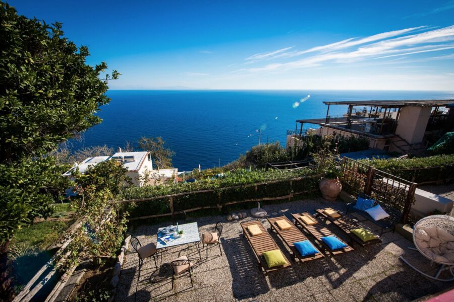 Coastal terrace with ocean view, lounging chairs, greenery, and vibrant cushions.