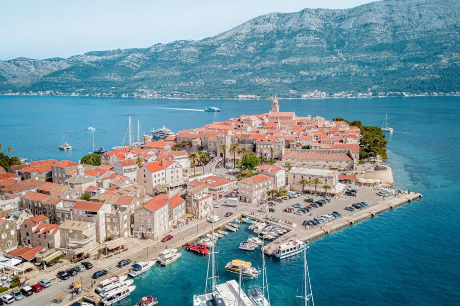 Coastal town with boats and mountains, featuring stone buildings and terracotta roofs under clear sky.