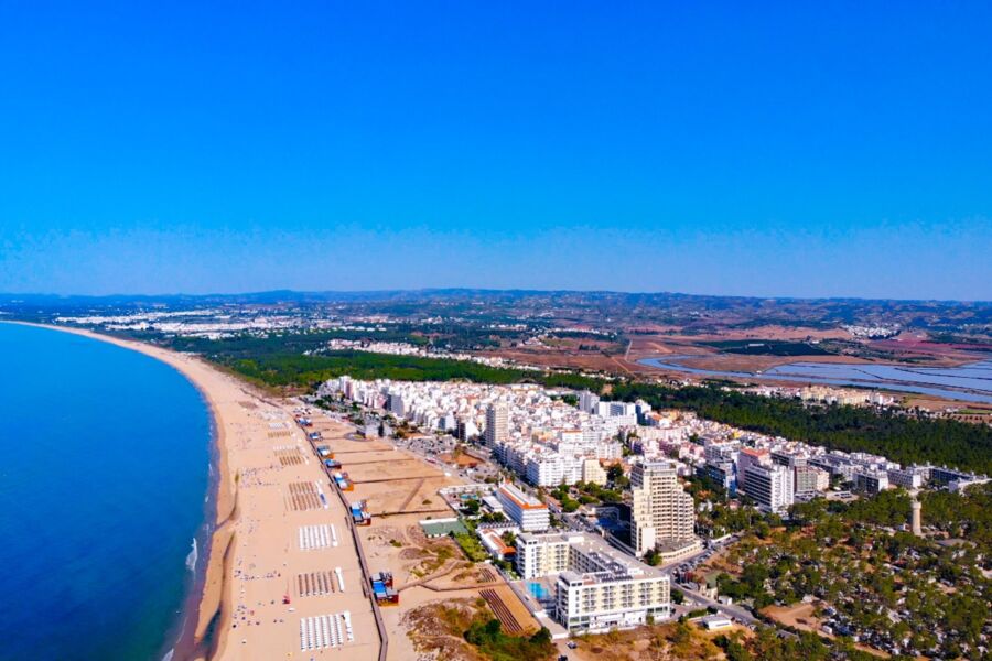Aerial view of a Monte Gordo coastal town with sandy beach, blue ocean, and whitewashed buildings.