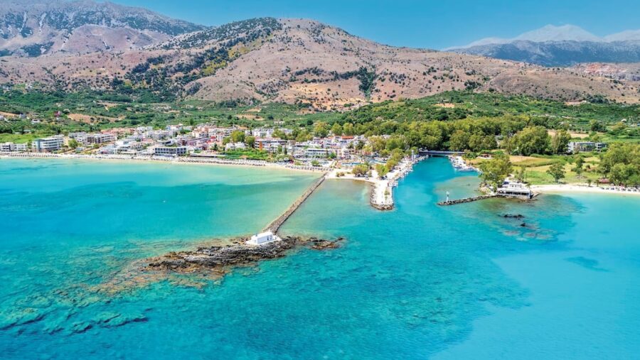 Aerial view of Mediterranean coastal town with beaches, turquoise sea, pier, and mountains.