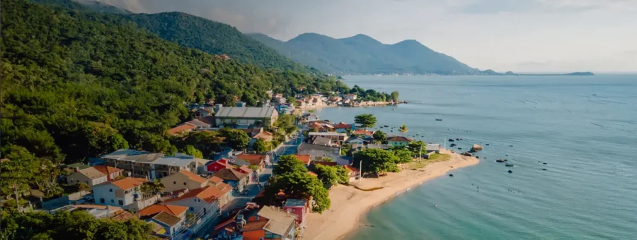 Aerial view of Ribeirão da Ilha coastal town with beach, fishing boats, and lush green mountains.