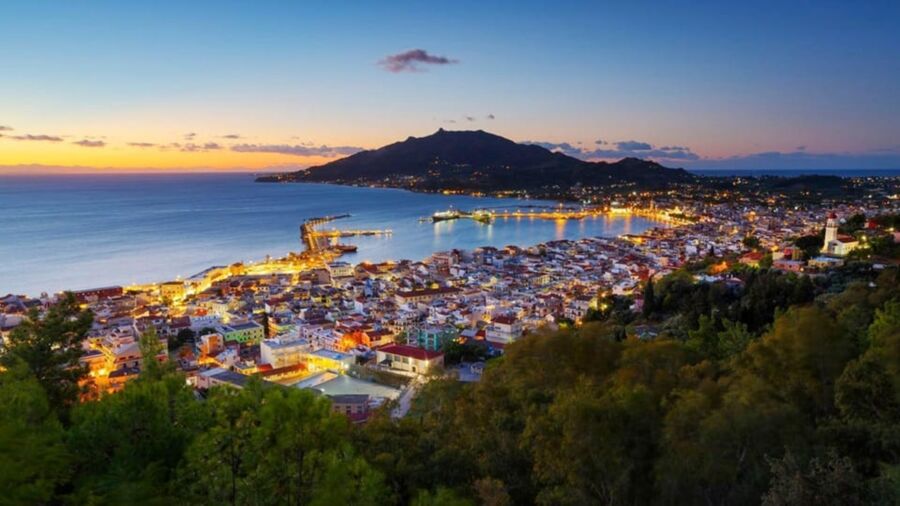 Dusk view of coastal town with sunset, mountains, Mediterranean architecture, harbor, and lighthouse.