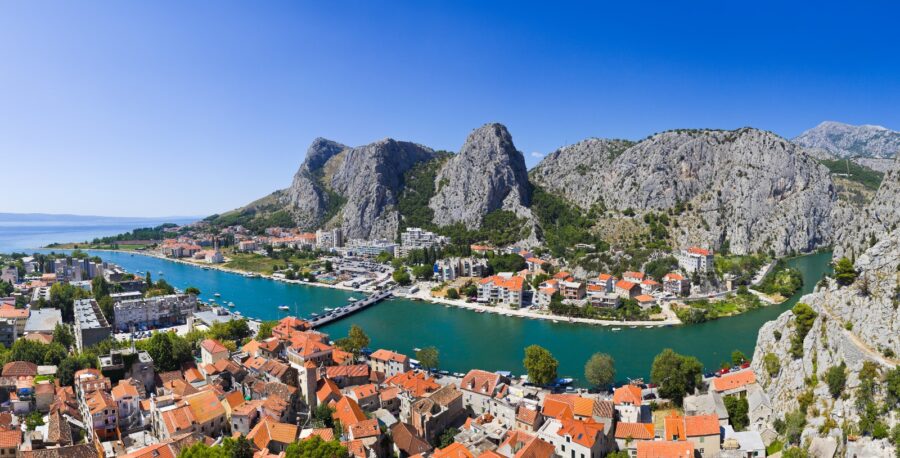 Scenic coastal town view with mountains, river, and vibrant architecture in sunlight.