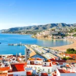 Picturesque coastal town with marina, colorful buildings, azure bay, lush greenery, hills, and sunset.