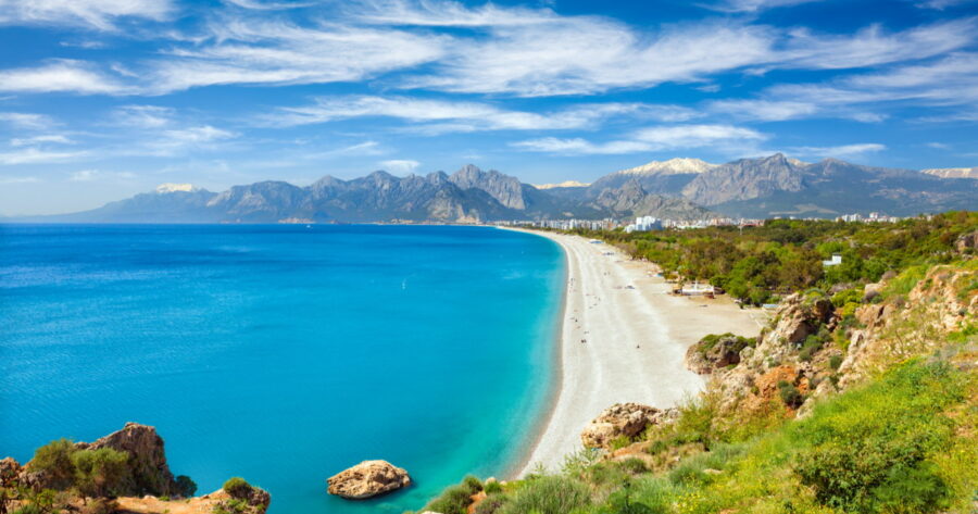 Turquoise Konyaaltı Beach coastline with pebbled shore, clear sea, resort city and snow-capped mountains.