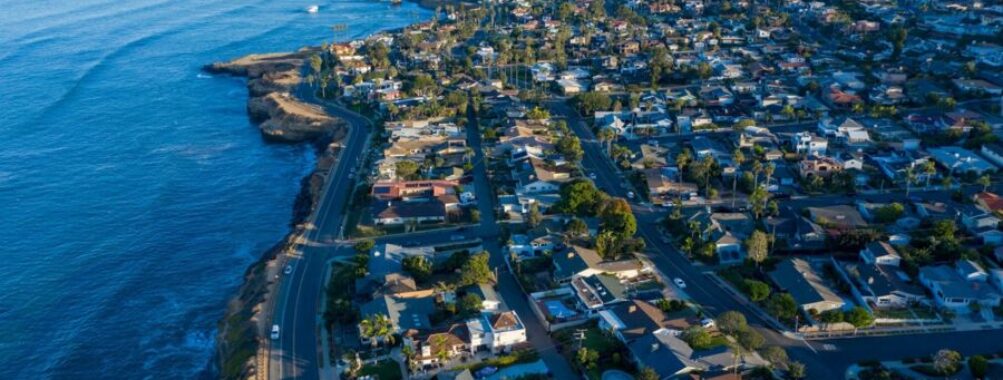Aerial view of coastal neighborhood with ocean, boats, diverse homes, and winding roads.