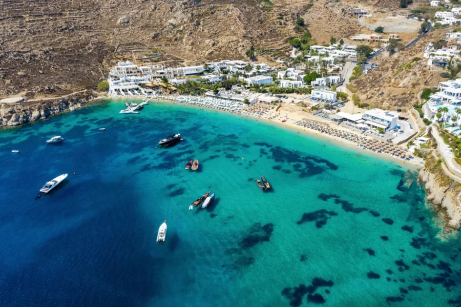 Aerial view of Psarou Beach: turquoise waters, yachts, sandy shore, resort buildings, hillside.