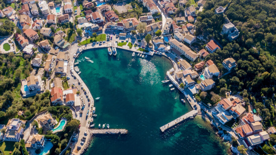 Aerial view of Kassiopi: coastal village, red-tiled roofs, turquoise bay, yachts, lush hills.