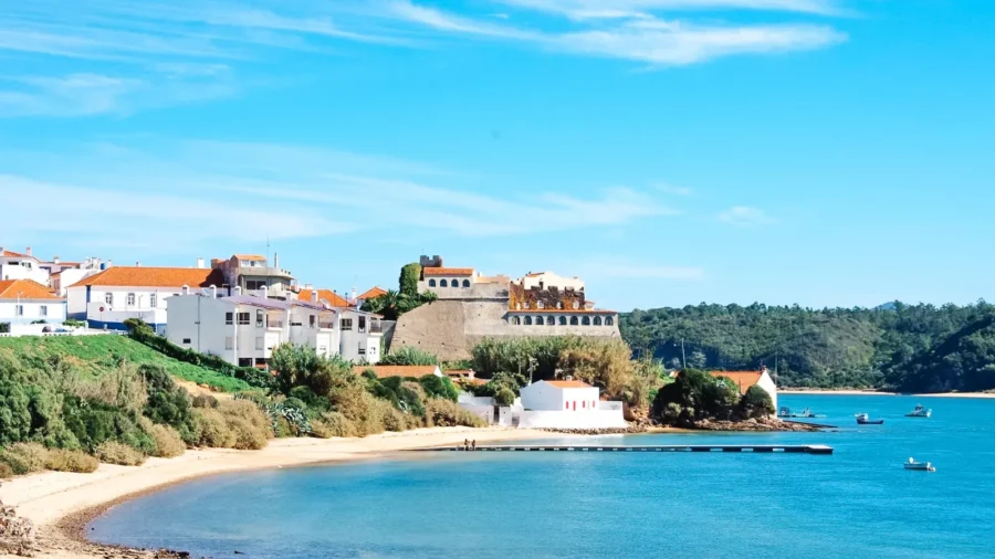 Coastal village beach with pier, turquoise water, white houses, terracotta roofs, and anchored boats.