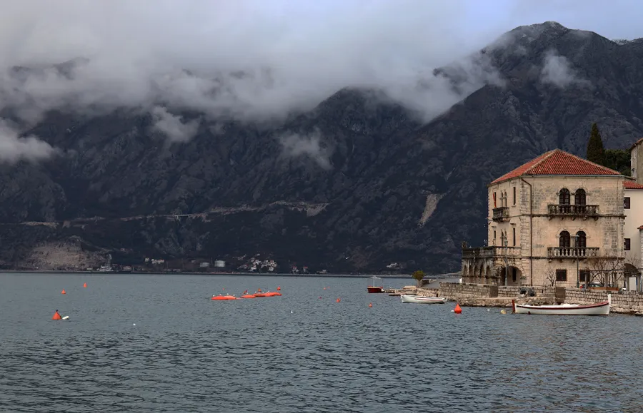 Stone coastal palace with red-tiled roof, moored boats and orange buoys against mountain backdrop.