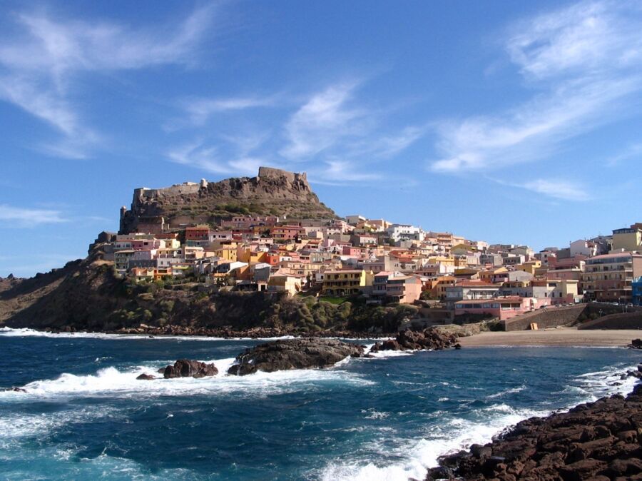 Picturesque coastal village with pastel buildings, rocky shoreline, and ancient fortifications under blue skies.