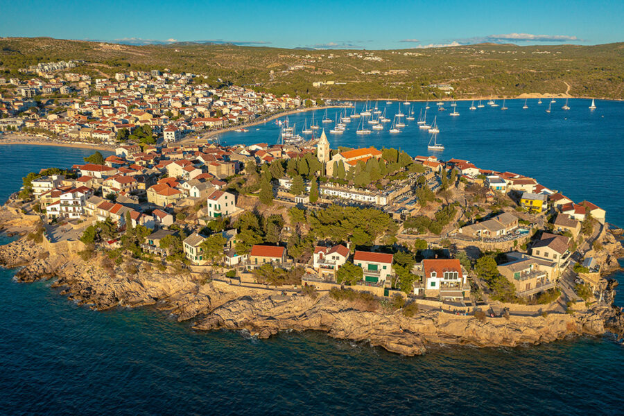 Scenic coastal town with Mediterranean architecture, boats, and lush hillsides under golden sunlight.