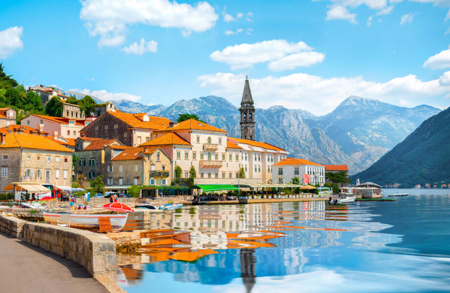 Charming Montenegro coastal village, mountains, boats, and clock tower in serene waterfront setting.