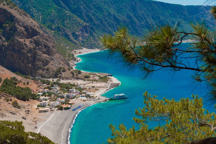 Coastal village with pine trees, serene bay, sandy beach, mountains, and sparkling blue water.